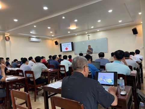 A group of people sitting at desks in a room with a person standing in front of a white boardDescription automatically generated